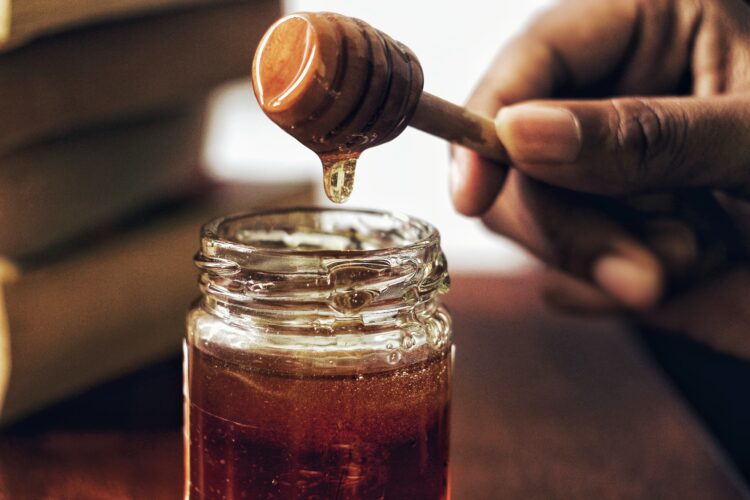 A hand holds a honey dipper above an open glass jar of honey, with honey slowly dripping from the dipper. Blurred books are visible in the background.