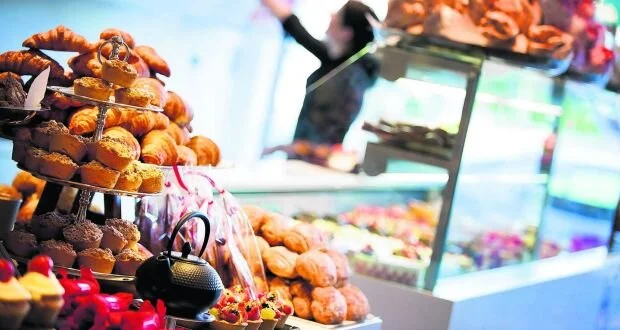A bakery display filled with assorted pastries, muffins, and croissants. A person is visible in the background behind a counter filled with more baked goods and colorful treats.