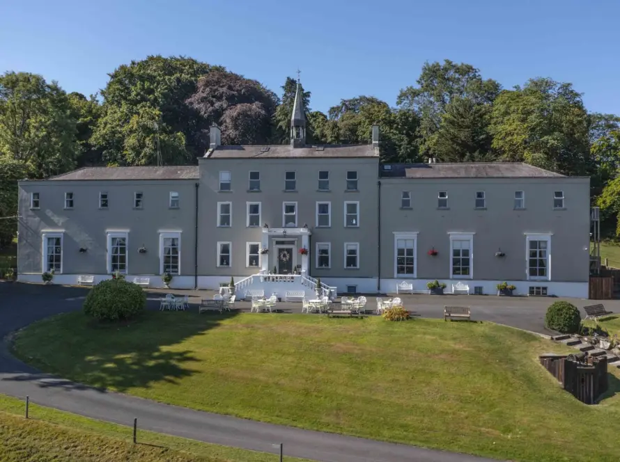 A large, symmetrical, gray manor house with white trim, surrounded by grass, trees, and outdoor seating areas on a sunny day. A curved driveway leads to the front steps and entrance.
