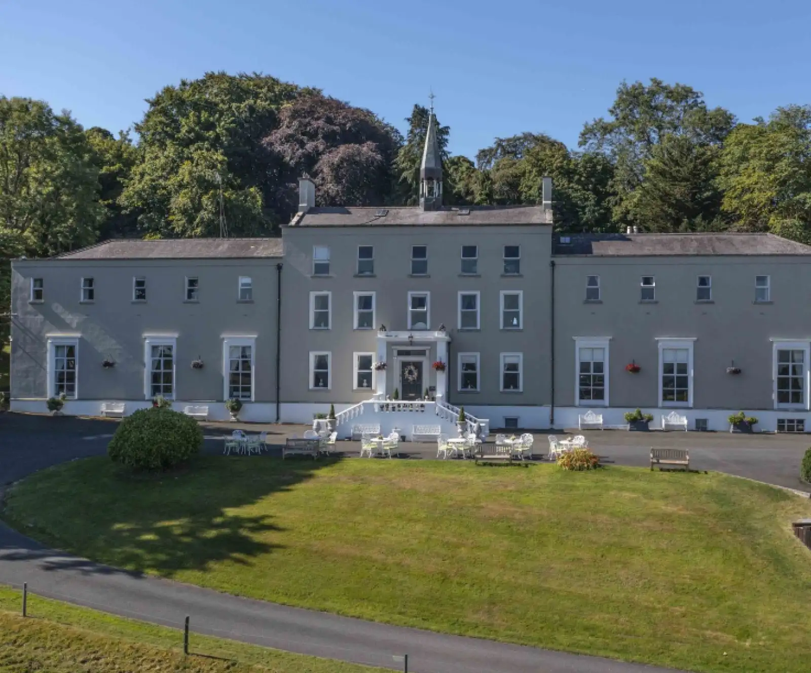 Large, light gray historic building with white trim, many windows, and a central entrance with steps. White outdoor furniture and benches are set on the green lawn, surrounded by trees under a clear blue sky.