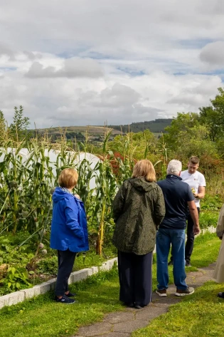 A group of people stand together on a garden path next to tall corn plants, with green hills and a cloudy sky in the background.