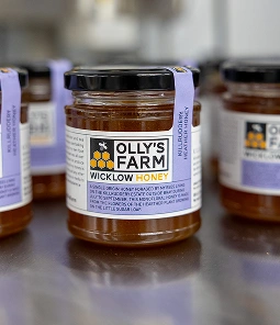 A jar of Olly's Farm Wicklow Honey with a purple label sits on a reflective surface; several more similar jars are blurred in the background.