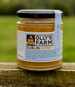 A jar of Olly’s Farm Dublin Honey sits on a wooden surface outdoors. The label features a honeycomb graphic and product description, with a blue ribbon on the lid. The honey inside is light golden in color.