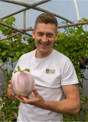 A smiling man in a white shirt stands in a greenhouse, holding a large, round, light purple eggplant with green leaves. Lush green plants fill the background.