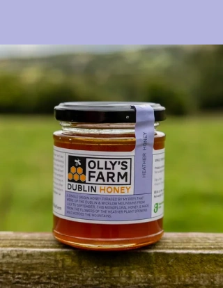A jar of Olly’s Farm Dublin Honey sits on a wooden surface outdoors, with a grassy field and trees blurred in the background. The label describes it as heather honey and highlights its origin and purity.