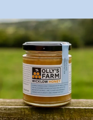 A jar of Olly's Farm Wicklow Honey with a blue seal sits on a wooden surface outdoors, with a blurred grassy background.